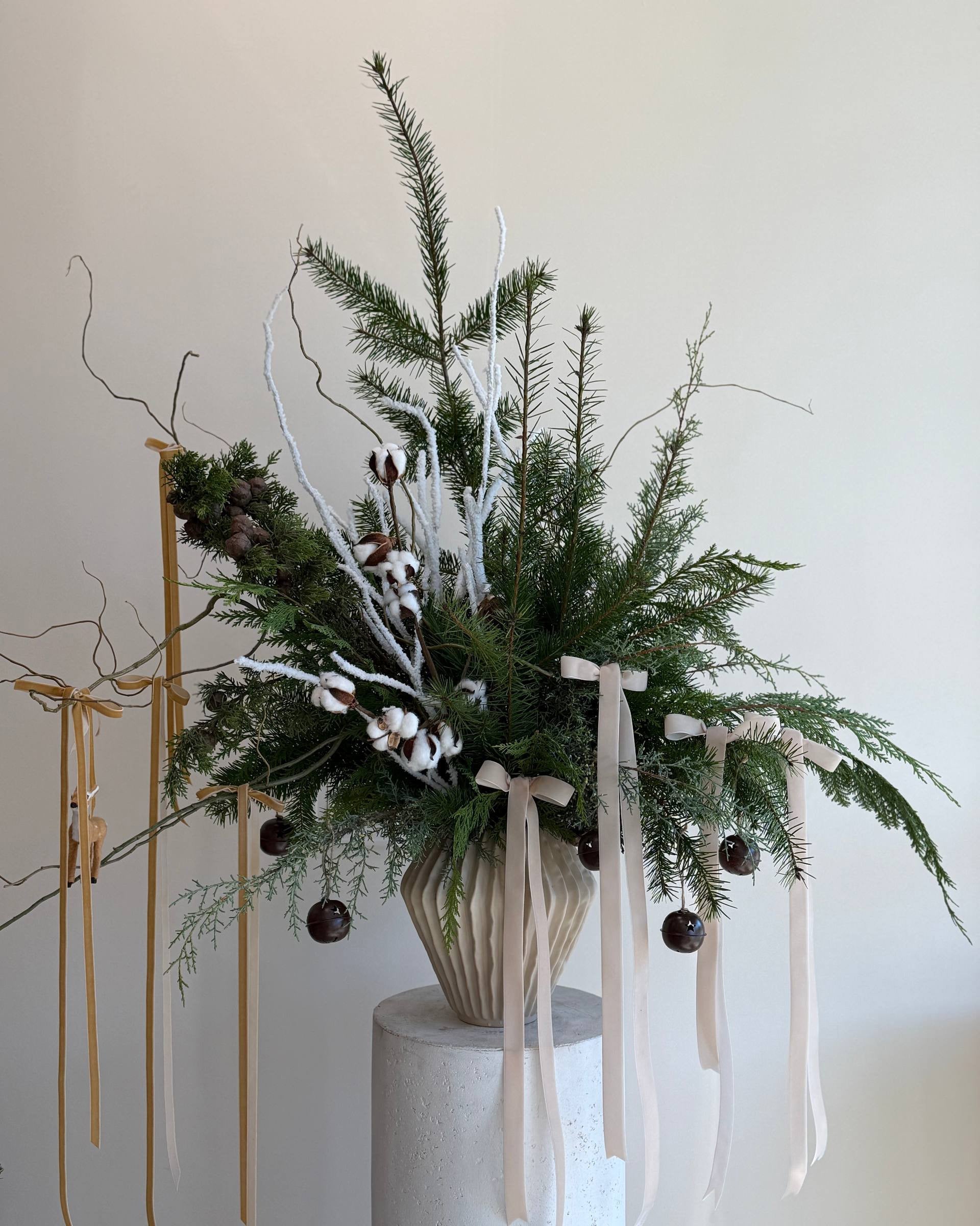 Decorative arrangement with greenery, ribbons, and small ornaments on a white pedestal against a light background.